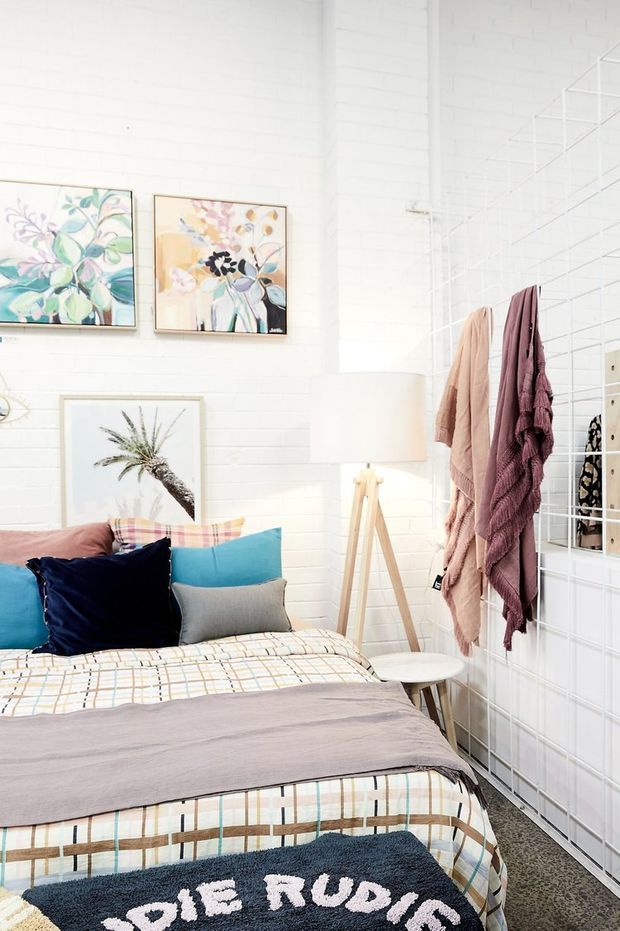 Bright, playful bedroom with a bed dressed in a patterned plaid duvet and layered turquoise, navy, grey, and blush cushions. White-painted brick walls display colourful botanical artwork, while a timber tripod floor lamp, small side table, and white wire-grid partition with draped throws add texture and personality.