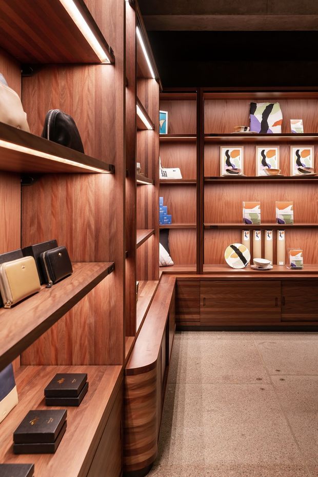 A warm, richly detailed retail-style display space lined with dark timber veneer shelving, cabinetry, and integrated linear lighting. Built-in shelves present books, artwork, small objects, handbags, and other merchandise, with a central timber-framed display wall and a polished neutral floor. The composition highlights bespoke joinery and a refined, gallery-like fit-out.