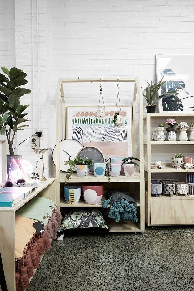 A bright, eclectic retail-style interior with white-painted brick walls, polished speckled concrete flooring, open timber display shelving, colourful ceramic vessels, textiles, plants, framed artwork, and a low upholstered bench. The space has a playful handcrafted feel with abundant merchandise and decorative objects.