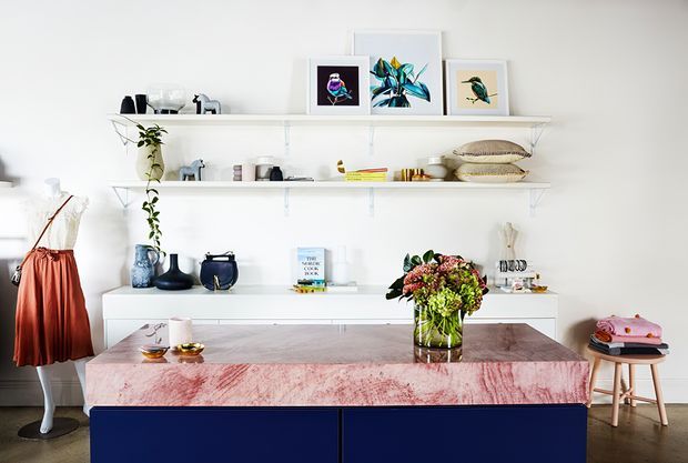 Bright, carefully styled dining area with a large dark blue built-in sideboard or island, a striking pink-and-white stone tabletop, open white display shelving, framed botanical artwork, ceramics, and a floral arrangement. A small stool and clothing display appear at the sides, while the dark timber floor adds contrast to the predominantly white interior.
