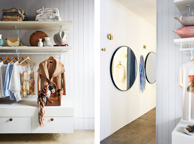 A bright, carefully organised walk-in wardrobe or dressing room with open shelving, hanging rails, drawers, folded clothing, accessories, and coats. White vertical wall panelling and cabinetry create a clean backdrop, while a round black-framed mirror and small wall-mounted hooks add functional and decorative detail. The space opens toward a light-coloured corridor or adjacent area with polished concrete flooring.