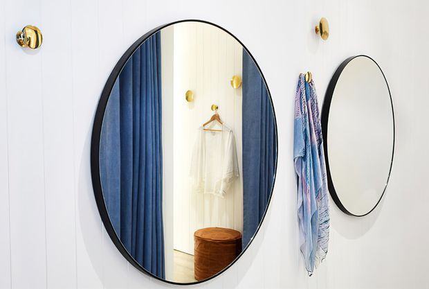 A compact dressing or wardrobe area with white vertical wall panelling, a large black-framed oval mirror, and a smaller round wall mirror. Deep blue curtains or fabric panels flank the oval mirror, reflecting a hanging white garment and a small brown upholstered stool. Gold wall hooks and a draped blue patterned textile add decorative accents.