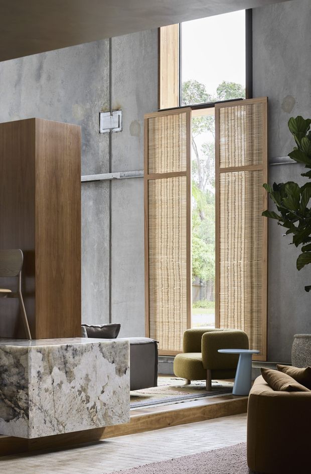 A refined contemporary living room with raw grey concrete walls, exposed conduit, tall timber-framed glazing, and woven timber blinds. The space features a large natural-stone-clad island or console, warm timber flooring and cabinetry, an olive-green lounge chair, a brown sofa, and layered neutral rugs. The mix of industrial finishes, organic materials, and sculptural furniture creates a warm, sophisticated atmosphere.