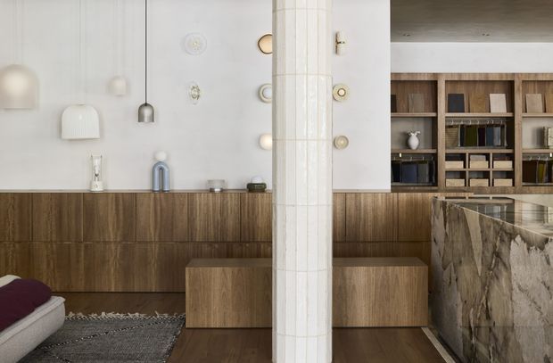 A refined open-plan interior combining a kitchen and living area. Warm timber cabinetry and flooring contrast with white walls, sculptural wall and pendant lighting, and a prominent vertically tiled structural column. A large veined stone island occupies the right side, while open timber shelving displays books and objects in the background. The image has a calm, sophisticated, contemporary feel.
