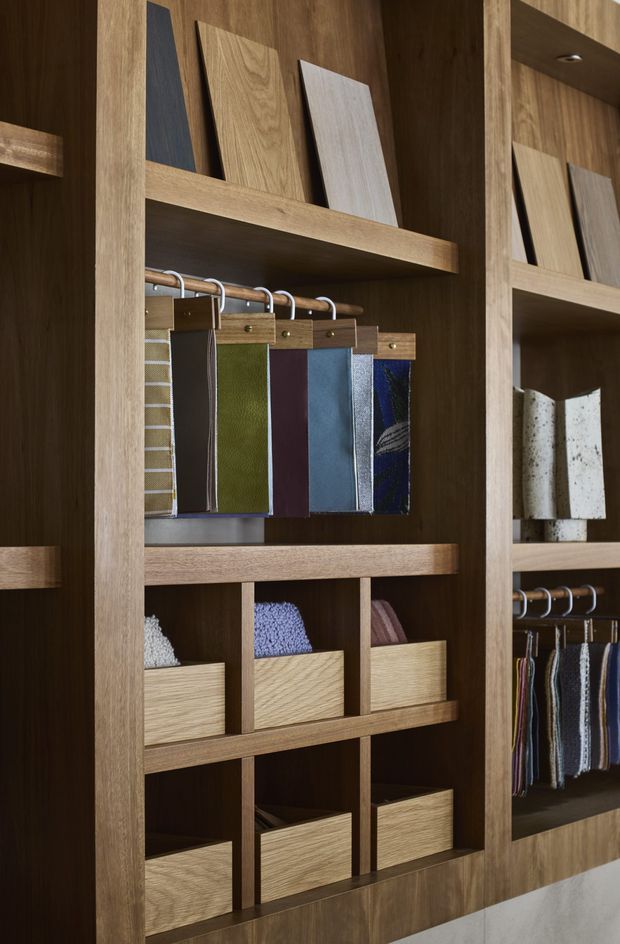 Close-up view of a custom timber-finished display and storage wall, with open shelving, cubby drawers, hanging fabric or material sample swatches, and upright wood and stone samples. The built-in joinery has a warm, refined showroom or design-studio appearance.