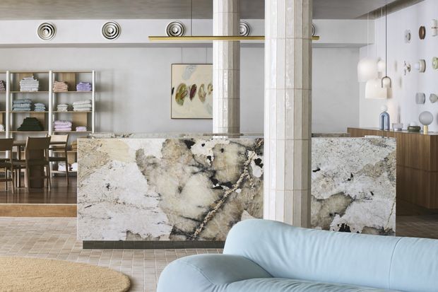 A refined open-plan interior featuring a dramatic veined stone island or counter, tiled structural columns, and a pale blue curved sofa in the foreground. A dining area with open shelving is visible to the left, while sculptural lighting, artwork, and timber cabinetry add understated character. The palette is light and neutral with natural stone, pale blue upholstery, and warm timber accents.