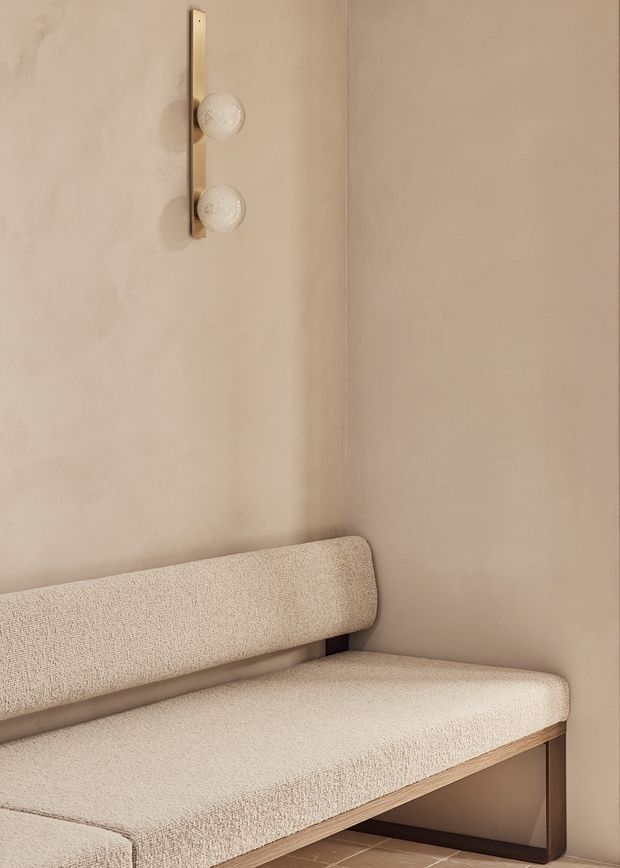 A close-up of a softly styled built-in or compact upholstered bench set into a warm beige plastered corner. The bench has cream textured upholstery, a rounded backrest, light timber edging, and dark slender supports. Above it is a minimalist vertical wall light with two frosted globe bulbs and a brass-toned backing. The overall composition is calm, monochromatic, and tactile.