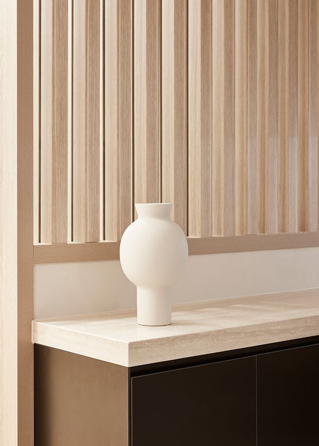 Close-up interior detail of a refined built-in console or cabinetry unit. The composition features vertical light timber slatted wall panelling, a pale stone or engineered-stone benchtop, and dark lower cabinetry, styled with a simple matte white sculptural vase. The image emphasizes warm natural materials, clean lines, and subtle tonal contrast.