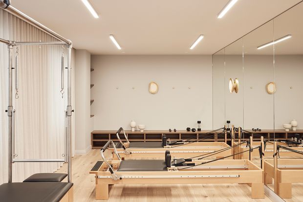 A bright, refined Pilates studio with multiple wooden Pilates reformer machines arranged in rows. The room has pale timber flooring, warm off-white walls and ceiling, full-height mirrored panels on one wall, linear recessed ceiling lights, and a curtained changing or treatment area at left. Minimal shelving and small decorative objects line the rear wall.
