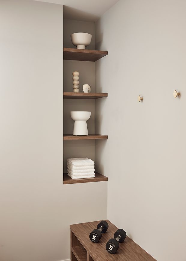 A minimalist built-in wall niche with four dark timber shelves displaying neutral ceramic vessels and folded white towels. Below, a timber storage bench or cabinet holds dumbbells, suggesting a compact home gym or wellness area. The space has warm beige plaster walls, simple brass wall hooks, and soft neutral styling.