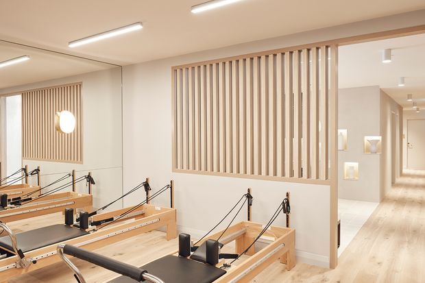 Bright, contemporary Pilates studio with several wooden reformer machines arranged in rows. The room features pale timber flooring, white walls and ceiling, linear LED lighting, a large wall mirror, and a decorative vertical timber batten screen separating the studio from a softly lit hallway with recessed display niches.