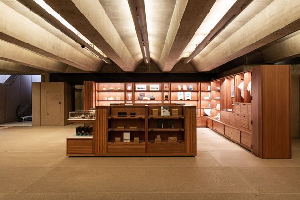 A warmly lit retail or showroom interior with extensive dark timber display cabinetry, central freestanding shelving, built-in wall shelving, and a dramatic exposed timber beam ceiling. The space has a refined, gallery-like atmosphere with neutral flooring and carefully integrated linear lighting.