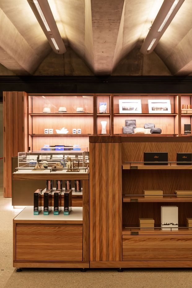 A warmly lit retail showroom or commercial display interior with richly finished timber cabinetry, glass display cases, open shelving, and curated product merchandise. The space features a dramatic exposed concrete coffered or ribbed ceiling with integrated linear lighting, dark framing around illuminated display niches, and a polished, high-end retail fit-out.