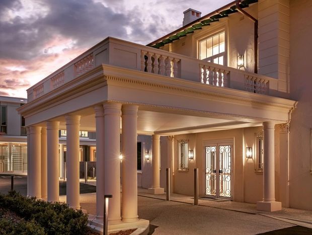 A grand classical-style building entrance at dusk, featuring a covered porte-cochère supported by tall round columns, ornate cornices, balustrades, pilasters, decorative wall lighting, and a glazed double entry door with wrought-iron detailing. The upper level has a terrace and warm interior lighting, while a dramatic cloudy sky forms the background.
