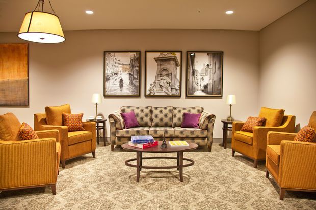 A warmly lit, symmetrical traditional living room with a tufted patterned sofa, multiple mustard-gold upholstered armchairs, a round wooden coffee table, framed architectural artwork, table lamps, and a large patterned area rug. The room has beige walls, recessed ceiling lights, and a pendant fixture.