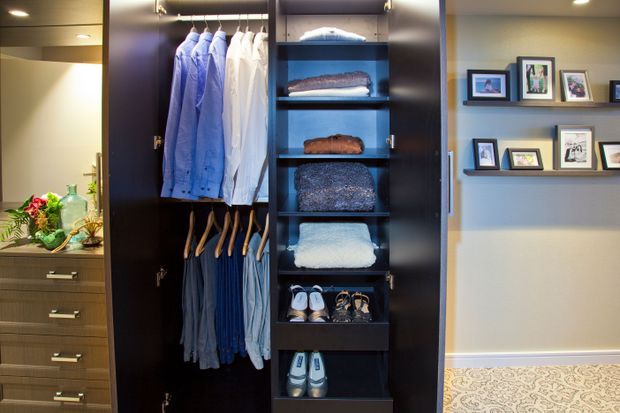 A custom built-in wardrobe and dressing area with dark timber cabinetry, hanging rails, open shelving for folded clothing, and shoe storage. A matching drawer unit with decorative items is visible to the left, while floating display shelves with framed photographs are mounted on the adjacent wall. The space has warm recessed lighting, neutral walls, and patterned carpet.