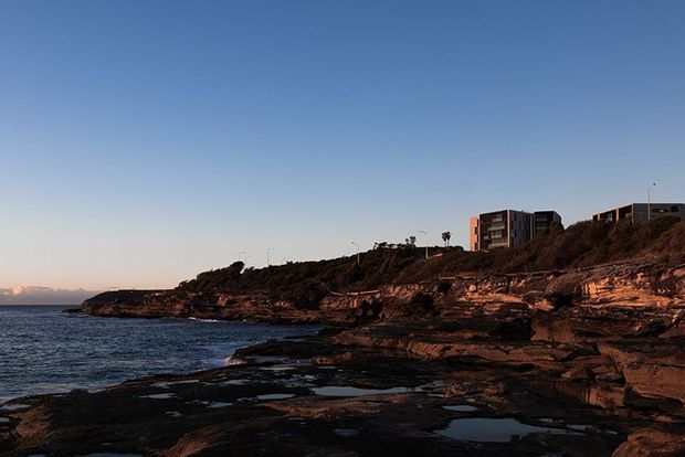 A wide coastal landscape at low sun, showing dark rocky tidal shelves and pools in the foreground, calm ocean to the left, and a rugged elevated cliff with sparse vegetation and modern residential buildings along the ridge. The scene has warm sunset highlights against a clear blue sky.