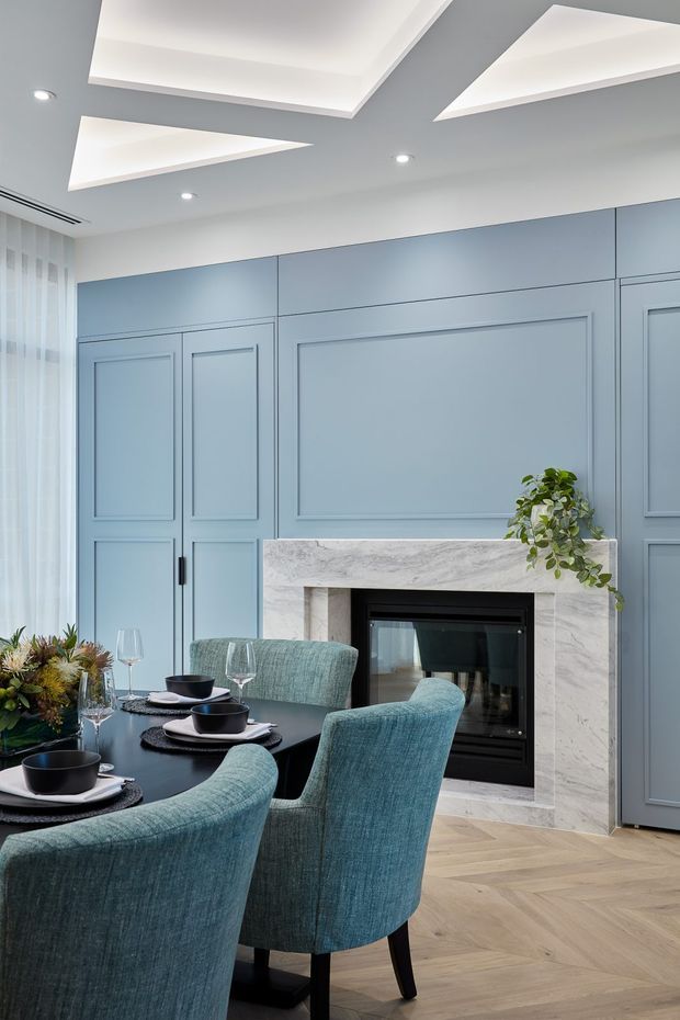 A refined dining area with a black dining table and upholstered teal-blue chairs, set against a blue-grey paneled wall with a built-in fireplace surrounded by pale veined stone. The room features light timber herringbone flooring, sheer curtains, recessed downlights, and dramatic geometric recessed ceiling details. Table settings, glassware, and a floral arrangement add a formal yet inviting atmosphere.