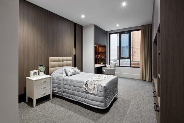 A contemporary bedroom with a grey upholstered bed and quilt, grey carpet, dark timber wall panelling and built-in wardrobe cabinetry. A white bedside table, small desk or vanity area, shelving, recessed lighting, and full-height window with curtains complete the refined, neutral interior.