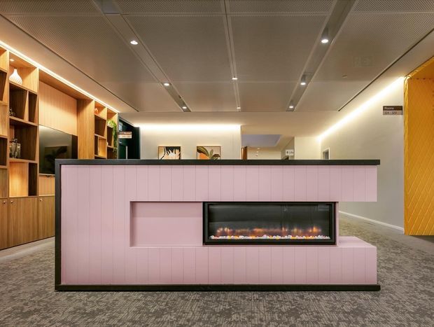 A contemporary commercial interior featuring a long pink reception desk or built-in counter with a linear electric fireplace inset into its front. Warm timber shelving and cabinetry line the left wall, while acoustic perforated ceiling panels, recessed lighting, grey carpet, and a yellow textured feature wall complete the space.