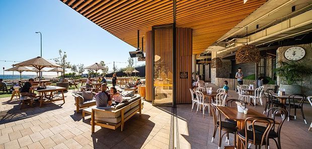 A bright outdoor dining and lounge area beside a waterfront, with timber picnic-style tables, upholstered lounge seating, umbrellas, and a covered restaurant terrace. The space features a prominent slatted timber ceiling, timber screening around a central column, exposed services, patterned masonry paving, and mixed café furniture. The setting is relaxed and contemporary with strong indoor-outdoor connection.