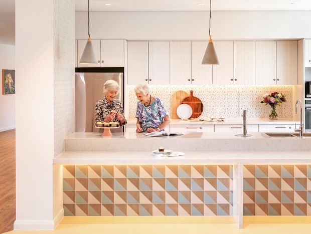 Bright contemporary kitchen with a large pale stone island, light cabinetry, terrazzo-style backsplash, stainless-steel appliances, and a distinctive geometric tile-clad island base in peach, cream, and muted blue. Two pendant lights hang over the island, with open shelving and kitchen joinery behind.