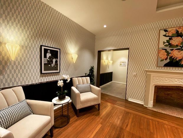 A refined traditional sitting room with two upholstered armchairs, patterned cushions, a small side table, decorative wall sconces, geometric wallpaper, timber flooring, framed artwork, and an ornate white fireplace with a timber-lined firebox. A large mirror reflects the room and additional lighting.