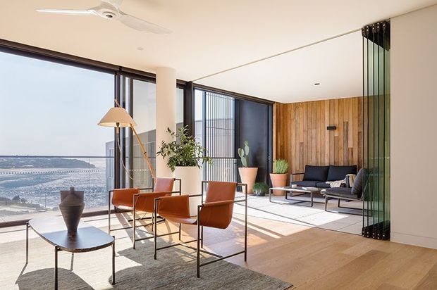 Bright, spacious contemporary living and dining area with floor-to-ceiling glazing overlooking a coastal urban landscape. The room features light timber flooring, a warm vertical timber feature wall, dark-framed glazing, a glass sliding partition, a minimalist dining table with leather chairs, and a separate lounge seating area. Natural light and a neutral palette create an airy, refined atmosphere.