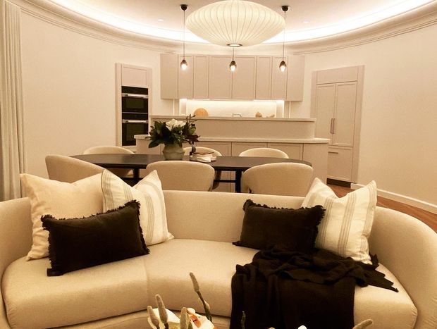 A warm, refined open-plan interior combining a living room, dining area, and compact kitchen. The foreground features a large cream upholstered sofa with contrasting dark cushions and throw, while a dark dining table with upholstered chairs sits behind it. The kitchen has pale built-in cabinetry, integrated appliances, a peninsula, and layered pendant lighting. Decorative wall mouldings, a curved ceiling with recessed lighting, and warm timber flooring add a polished traditional character.