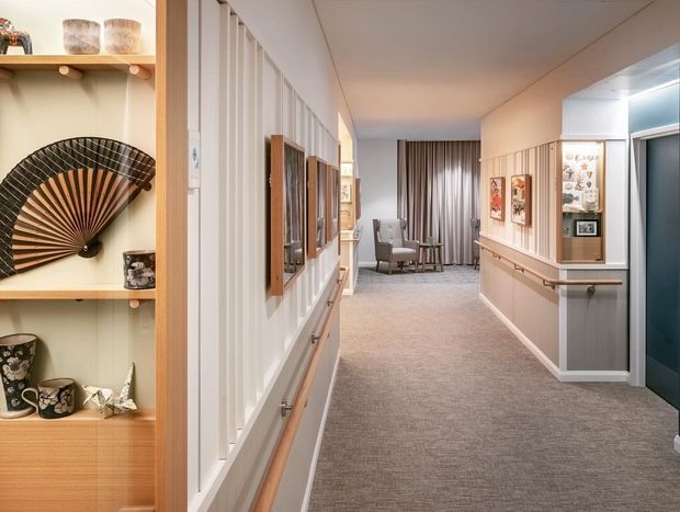 A long, softly lit residential hallway with grey carpet, white vertical wall panelling, timber handrails, and warm timber display shelving. Framed artwork and decorative objects line the corridor, with a small seating area visible at the far end and a dark blue doorway to the right.