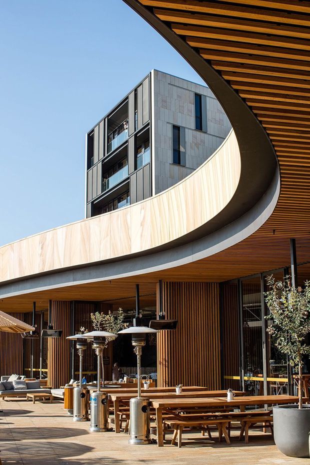 A contemporary multi-storey building with a prominent sweeping curved balcony or canopy, grey stone-clad upper facade, dark metal framing, and extensive warm timber soffit and vertical timber screening. The ground level forms an outdoor hospitality terrace with timber picnic-style tables, lounge seating, heaters, umbrellas, potted trees, and glazed openings beneath the curved structure. Clear blue sky and strong sunlight emphasize the sculptural architectural form.