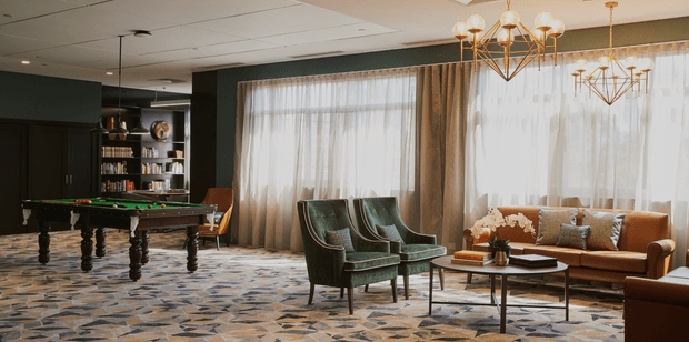 A refined lounge and billiards room with dark green walls, large sheer-curtained windows, patterned carpet, a traditional pool table, velvet lounge chairs, a cognac leather sofa, and geometric brass pendant chandeliers. The space combines vintage-inspired furnishings with contemporary hospitality-style detailing.