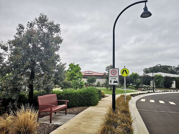 A landscaped pedestrian walkway in a residential or community setting, with a red bench, ornamental grasses, hedges, mature trees, curved black street lighting, and a clearly marked 10 km/h shared zone beside a curved roadway and pedestrian crossing. Low-rise homes are visible in the background under an overcast sky.