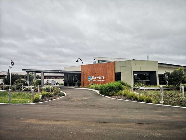 Street-level exterior view of a contemporary, low-rise commercial facility with a broad flat roof and covered entrance canopy. The facade combines warm vertical timber cladding, pale rectangular panel cladding, dark-framed glazing, and landscaped planting beds around a curved driveway. Signage identifies the building as Barwarre Community Centre.