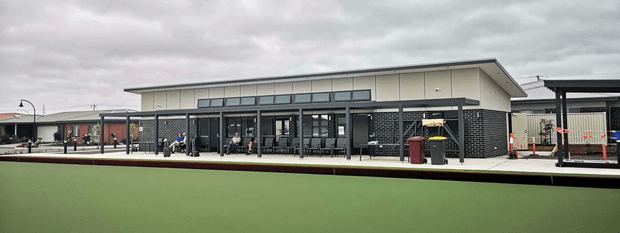 A low, contemporary single-storey building viewed from across a green artificial bowling or sports surface. The building has a broad shallow-pitched roof, dark brick masonry walls, extensive dark-framed glazed doors and windows, and a covered outdoor veranda or entry zone. Adjacent covered structures and a paved forecourt are visible, with an overcast sky and minimal landscaping.
