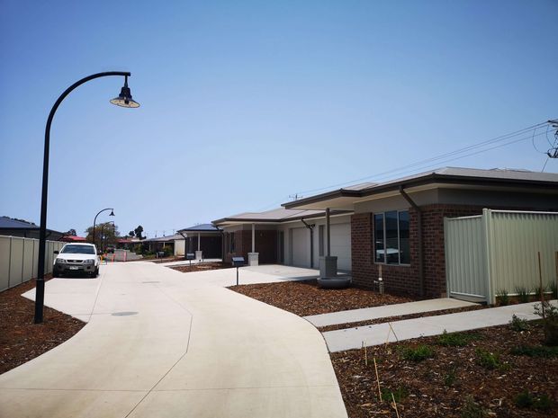 A suburban residential development viewed from a broad curved concrete driveway or shared access lane. The scene shows several single-storey homes with low-pitched hipped roofs, brick and rendered facades, covered porches, narrow front paths, fenced yards, and distinctive curved black street lamps. The landscaping is sparse and newly established, with gravel or mulch beds and young plants under a clear blue sky.