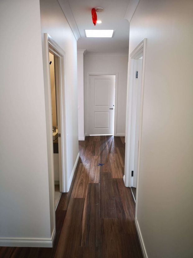 A narrow, bright residential hallway with white painted walls, decorative white door and window trims, multiple doorways, a rectangular skylight, and dark warm-toned timber flooring. A bathroom or utility space is partially visible through the left doorway. The corridor has a clean, traditional interior character with natural light from above.