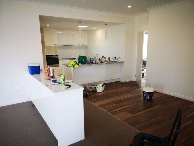 A bright, mostly white open-plan kitchen viewed from an adjoining carpeted living area. The kitchen has white flat-panel cabinetry, a long peninsula with a dark benchtop, integrated oven and cooktop, and dark timber-look flooring. The room appears unfinished or recently renovated, with cleaning supplies and miscellaneous items on the counters and floor. A doorway opens to another room on the right.