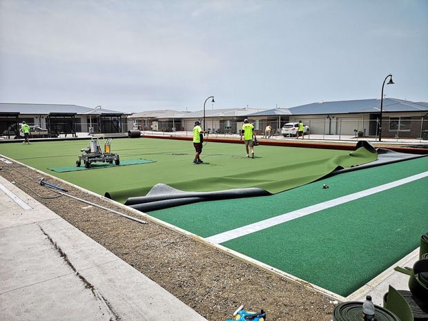 Outdoor sports facility under construction, showing multiple green synthetic playing surfaces with white boundary lines, partially rolled turf, installation equipment, workers in high-visibility clothing, perimeter fencing, and low modern buildings in the background.