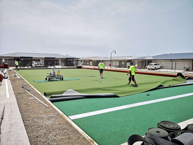 An outdoor lawn bowls facility under construction, with green synthetic bowling surfaces, white boundary markings, rolled turf materials, maintenance equipment, and several workers installing the playing surface. Low single-storey buildings and covered structures surround the courts.