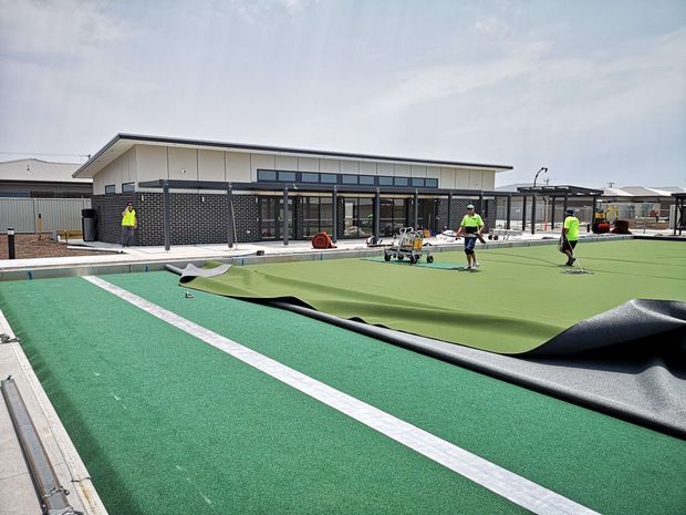 Exterior construction scene showing a low, modern community or sports facility beside a large outdoor artificial-turf playing surface. The green sports surface is partially lifted, exposing dark underlay, with white court markings and several workers and maintenance equipment visible. The building has a shallow single-pitched roof, extensive glazing, dark masonry walls, and covered outdoor areas.