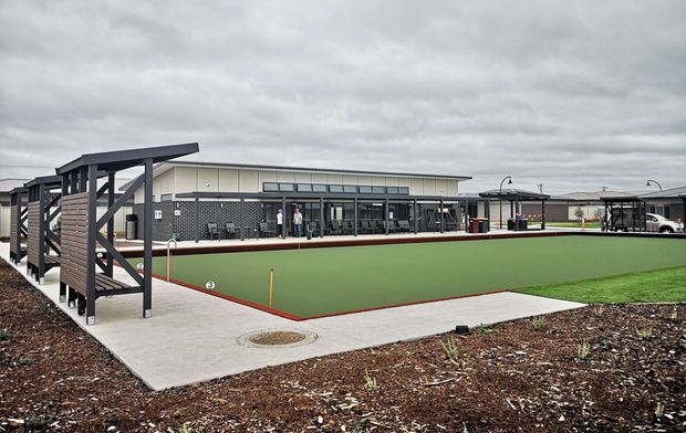 An exterior view of a contemporary low-rise community or sports facility, dominated by a large green synthetic bowling or sports court with red perimeter edging. The court is bordered by a concrete walkway and dark timber-and-metal screening structures, with a long glazed building containing covered seating or activity areas behind it. The scene is overcast and landscaped with sparse planting beds.