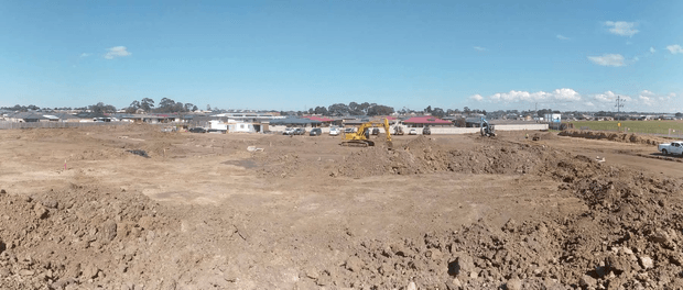A broad undeveloped construction site with freshly excavated and graded earth in the foreground. Several excavators, trucks, and construction vehicles are visible in the middle distance, with low-rise residential buildings and open fields beyond under a clear blue sky.