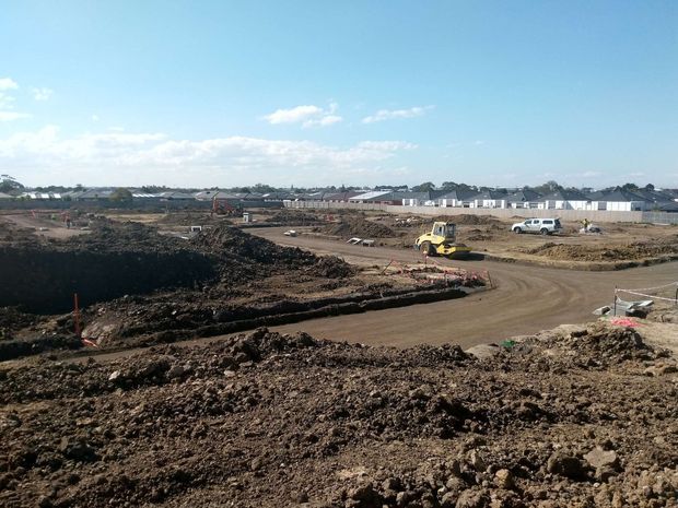 A broad construction site with freshly excavated and graded earth, dirt access roads, construction machinery, temporary fencing, and rows of newly built low-rise homes in the background under a clear sky.