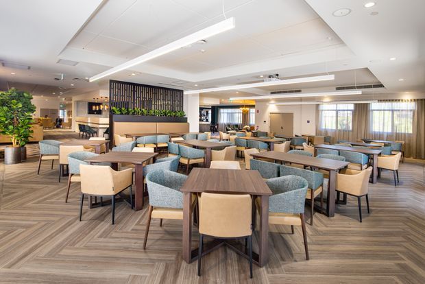 A spacious contemporary dining and hospitality interior with numerous square and rectangular tables, upholstered chairs, a central planted timber-slat divider, and a service counter in the background. The room features patterned wood-look flooring, a white suspended ceiling with linear pendant lighting and recessed downlights, and large curtained windows.