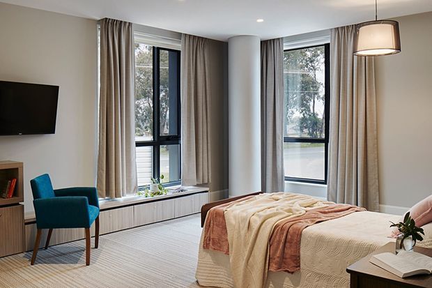 A bright, refined bedroom with a large bed dressed in layered cream and blush textiles, teal upholstered armchair, wall-mounted television, built-in window seat or low joinery, floor-to-ceiling curtains, and tall black-framed corner windows. A cylindrical column and contemporary pendant light add architectural interest. The room overlooks greenery and appears calm and spacious.