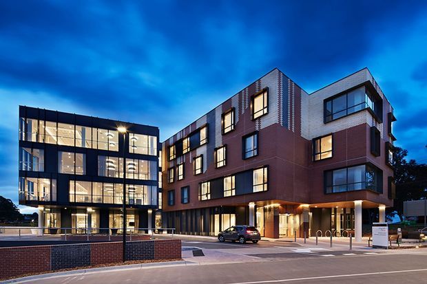 A dusk exterior photograph of a contemporary multi-storey commercial or institutional building complex. The composition shows a dark glazed rectilinear wing beside a larger red-brown brick building with projecting sections, varied window openings, recessed ground-floor entrances, illuminated interiors, street frontage, bicycle racks, and paved public forecourt.