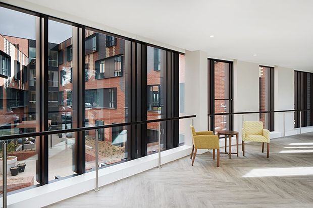 A bright contemporary interior corridor or communal lounge with floor-to-ceiling black-framed glazing overlooking a red-brick urban courtyard building. The space has a pale timber-look floor, white walls and ceiling, a slim black handrail, and a small seating arrangement of mustard and cream armchairs with a wooden side table. Strong natural light creates a clean, airy atmosphere.