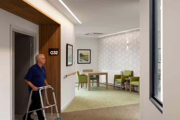 A contemporary residential or care setting hallway opening into a small dining and sitting area. The foreground features a white corridor with a warm timber-lined doorway surround, integrated linear ceiling lighting, a handrail, and a room sign. Beyond is a softly furnished space with a timber dining table, green upholstered chairs, pale patterned wallpaper, framed artwork, and carpeted flooring. A person using a walking frame is present, suggesting an accessible or aged-care environment.