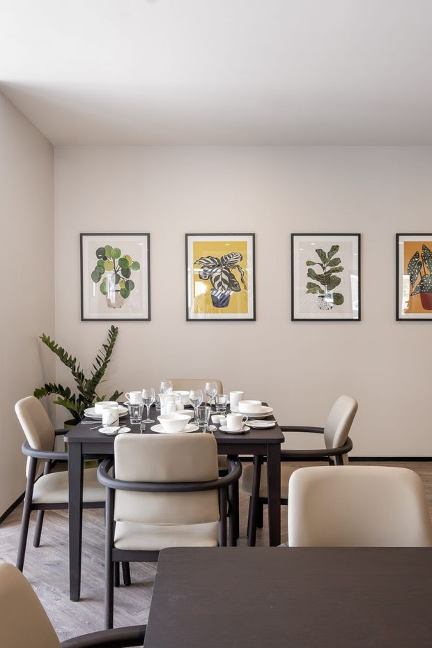 A refined dining room with a dark rectangular dining table set with white crockery and numerous wine and water glasses. Upholstered cream dining chairs with dark timber or black frames surround the table. The room has warm light-grey walls, a white ceiling, dark timber flooring, and a row of framed botanical artwork above the dining area. A potted plant adds greenery, while a second dark tabletop is visible in the foreground.